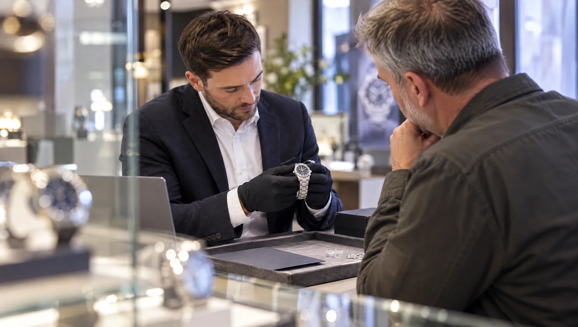 Watch dealer inspecting a pre-owned watch with a customer at the counter.